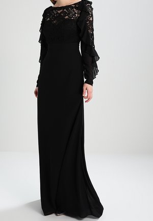 Femme portant une longue robe noire avec un corsage en dentelle et des manches longues transparentes à volants, debout devant un fond clair uni.