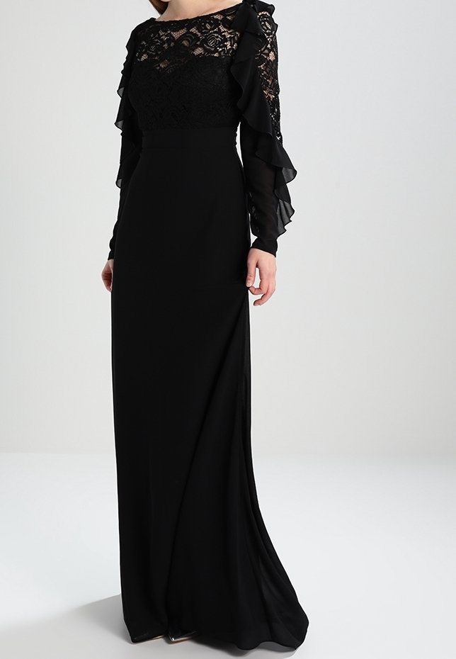 Femme portant une longue robe noire avec un corsage en dentelle et des manches longues transparentes à volants, debout devant un fond clair uni.