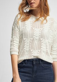 Lekki, dziany sweter w kremowym kolorze z teksturowanym wzorem. Posiada dekolt w kształcie łódki oraz zwinięte rękawy o długości do łokcia, zestawiony z ciemnoniebieskimi dżinsami.