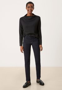 Haut noir à manches longues avec un col haut, associé à un jean skinny bleu foncé et des chaussures noires brillantes, se tenant devant un fond clair.