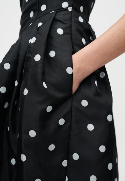 Svart klänning i tyg med stora vita prickar, med en sidoficka och veck för extra textur och dimension.