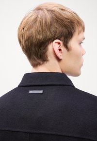 Manteau en laine noir avec une finition texturée, doté d'un col et d'une étiquette rectangulaire portant "Calvin Klein" au dos du cou.