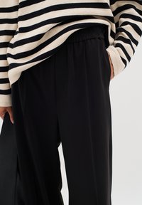 Schwarze Hosen mit lockerer Passform, ausgestattet mit einem gerafften Elastikbund. Gestreiftes Strickoberteil in Creme und Schwarz, das über die Hose fällt.