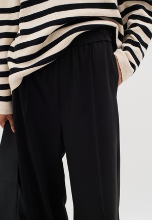Black trousers with a relaxed fit, featuring a gathered elastic waistband. Striped knit top in cream and black drapes over the pants.