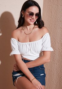 Hvid off-the-shoulder top med tekstureret stof, korte flæser på ærmerne, parret med mørk denimshorts med franske kanter og lommer.