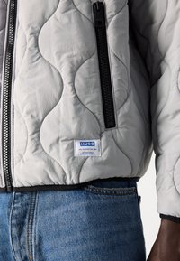 Quilted gray jacket with black zipper, ribbed cuffs, and HUGO label. Features a smooth texture and curved seam design. Worn over denim.