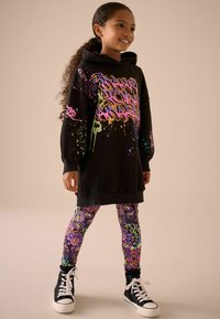 Schwarzer Hoodie mit bunten Graffiti-Designs, kombiniert mit gemusterten Leggings, die lebendige Formen zeigen. Schwarze High-Top-Sneaker runden das Outfit ab.
