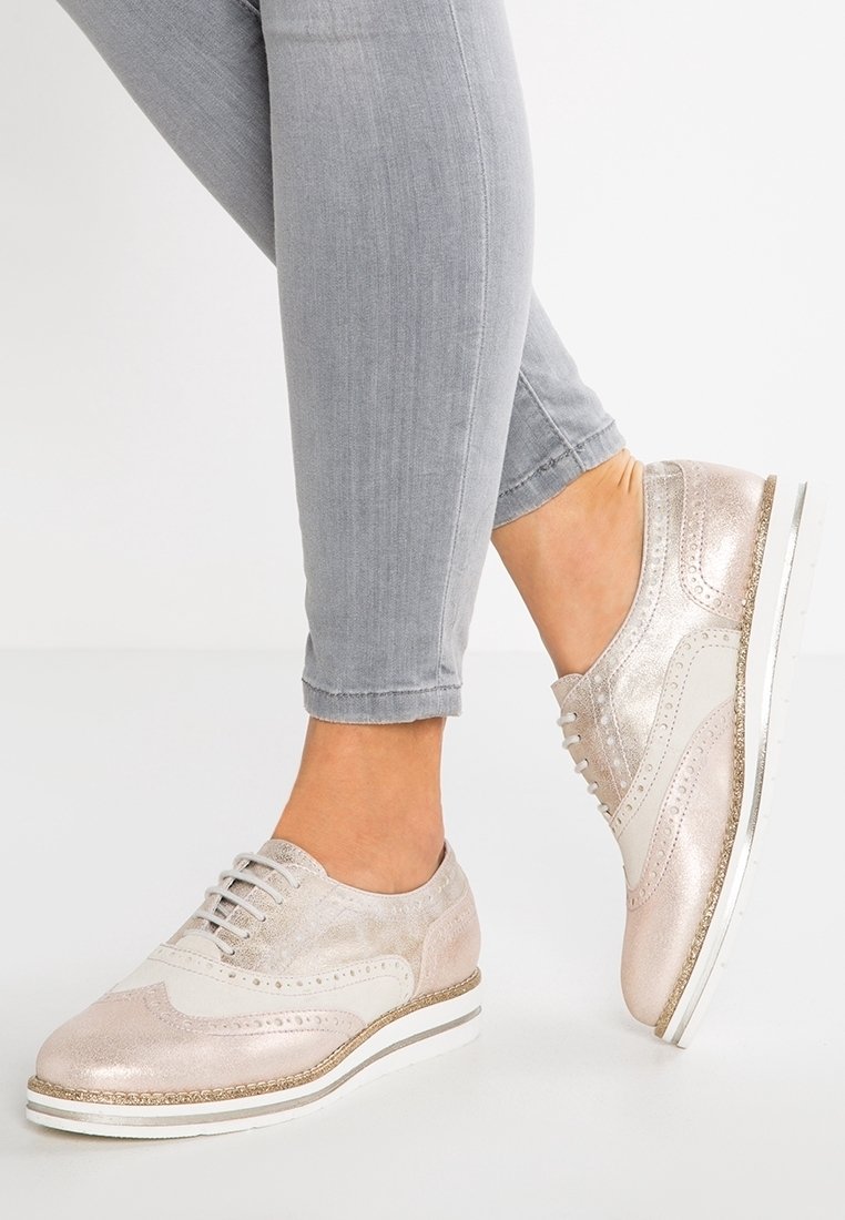 Pieds portant un pantalon slim gris clair et des chaussures à lacets bicolores rose pâle brillant et blanc avec des semelles blanches.