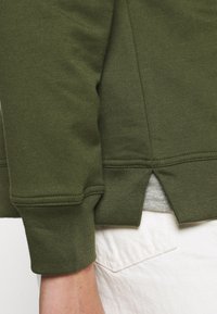 Sudadera verde oliva con una textura sutil, que presenta un detalle de dobladillo partido y puños acanalados, combinada con pantalones de color claro.