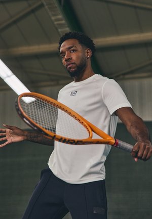 Camiseta deportiva blanca con logo en relieve, combinada con pantalones oscuros. Sostiene una raqueta de tenis naranja con un patrón de cuerdas negro en un entorno deportivo.