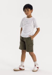 T-shirt bianco a maniche corte con testo verde, pantaloni cargo oliva con tasche laterali e sneakers bianche con accenti rossi e suole beige.