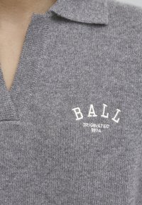 Polo de lana gris con cuello en V, tejido texturizado y un logo bordado en blanco que dice "BALL" con "ORIGINATED 1974" debajo.