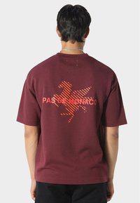 Mies, joka käyttää viininpunaista t-paitaa, jossa on oranssiraidallinen siivekkään hevosen grafiikka ja "PAS DE MONACO" -teksti selässä, mustia housuja ja hopeaketjukaulaketjua.