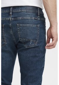 Los jeans de mezclilla azul oscuro presentan un diseño de cinco bolsillos con costuras visibles. La tela muestra una sutil textura y un leve desgaste en el bolsillo trasero.