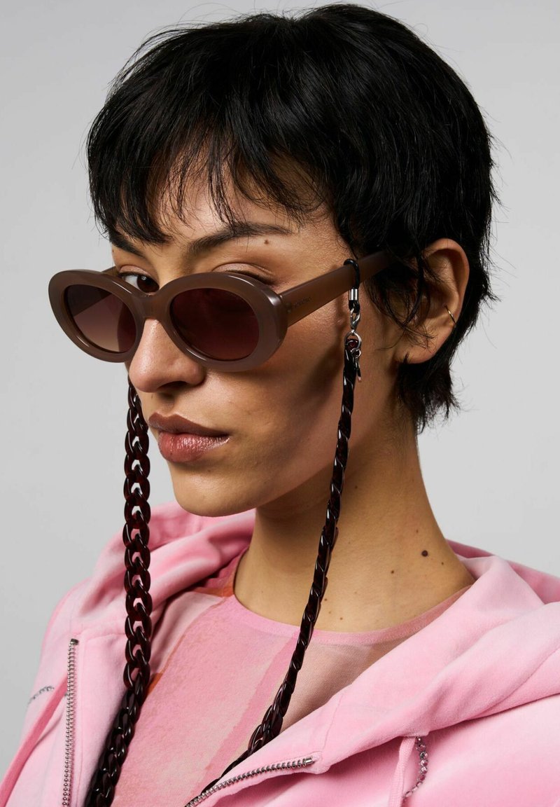 Bruna ovala solglasögon med en slät båge, mörka linser och en flätad svart kedja. Bärs med en rosa hoodie över en rosa topp.