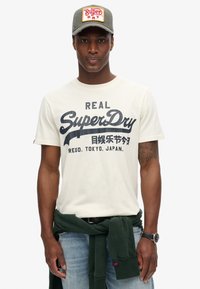 Camiseta de manga corta color crema con texto azul oscuro, que presenta la marca "SuperDry". Combinada con un sudadera verde atada a la cintura.