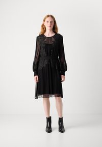 Robe plissée noire avec des détails en dentelle florale, des manches longues transparentes et une ceinture. Associée à des bottines noires. Texture lisse et fluide.