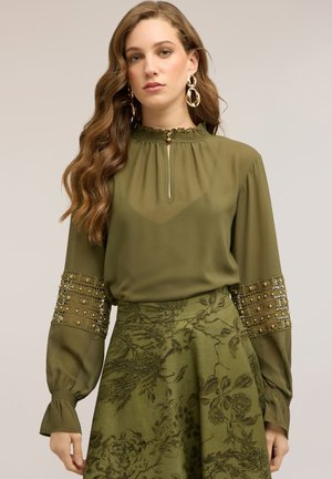 Nainen pukeutuneena oliivinvihreään puseroon, jossa helmiäiskoristellut pitsihihat, ja samanlaiseen kukkakuviolliseen hameeseen, poseeraa neutraalia taustaa vasten.