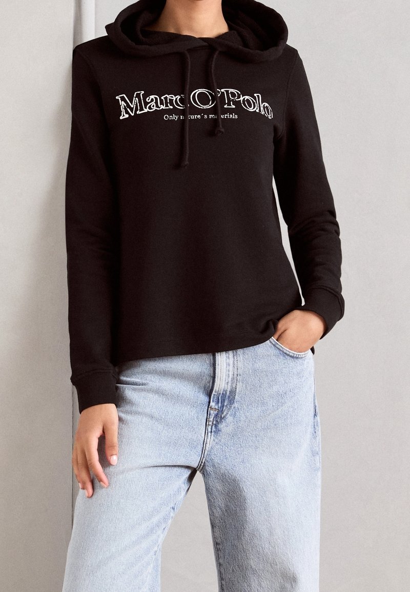 Sudadera negra con capucha y cordón ajustable, con logo frontal. Material de algodón, corte relajado, mangas largas, combinada con jeans de mezclilla azul claro.