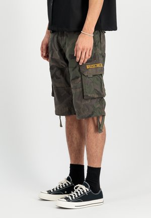 Mand iført mørke camouflage cargoshorts med en gul lap, sort skjorte, sorte sokker og sorte lave sneakers med hvide snørebånd.