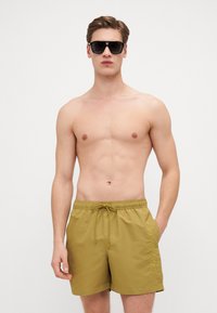 Shorts de bain pour homme en olive avec une taille à cordon de serrage et des poches latérales, présentant un logo en relief sur le côté gauche. Matériau lisse et léger.