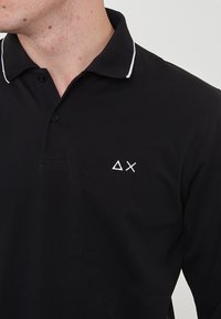 Polo nero in cotone con pattina a due bottoni e colletto con striscia bianca. Presenta un piccolo logo geometrico bianco sul lato sinistro del petto.
