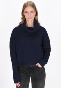 Vrouw in een marineblauwe coltrui met lange mouwen en zwarte jeans, staand met een hand in de zak tegen een witte achtergrond.