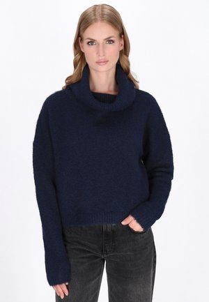 Vrouw in een marineblauwe coltrui met lange mouwen en zwarte jeans, staand met een hand in de zak tegen een witte achtergrond.