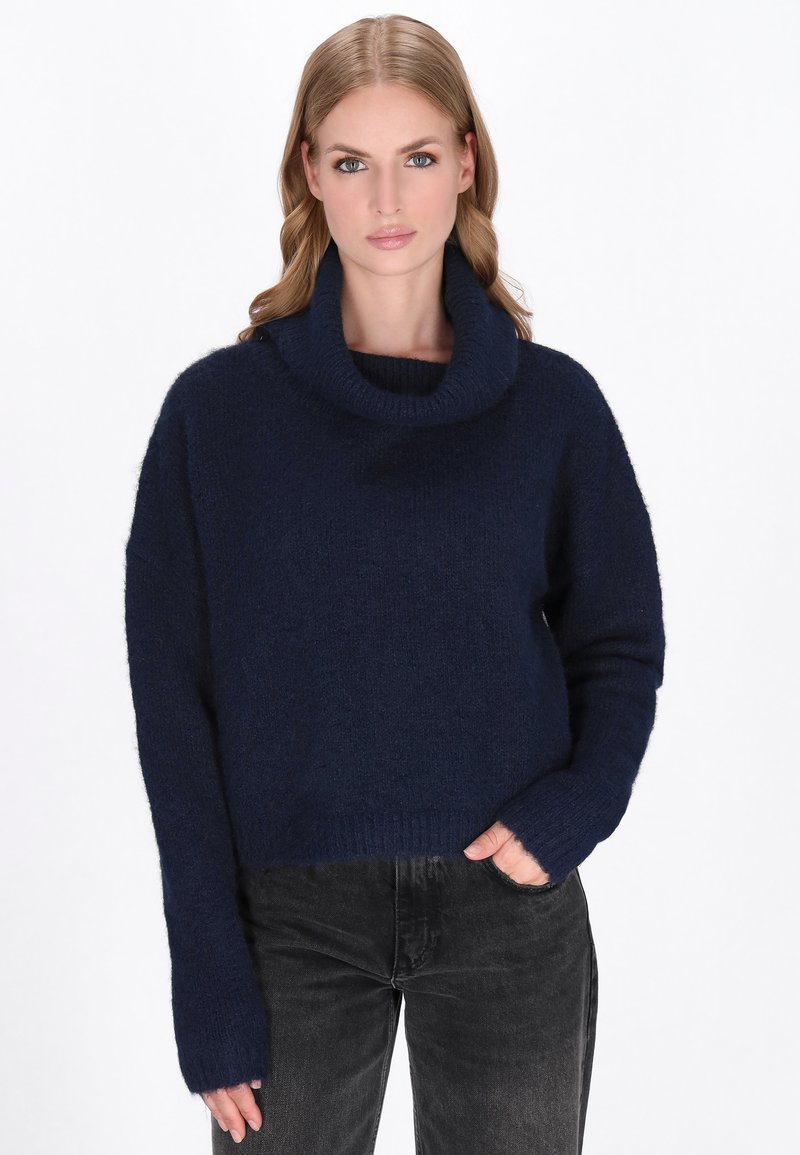 Donna che indossa un maglione blu navy con colletto a imbuto e maniche lunghe, con jeans neri, in piedi con una mano in tasca su uno sfondo bianco.