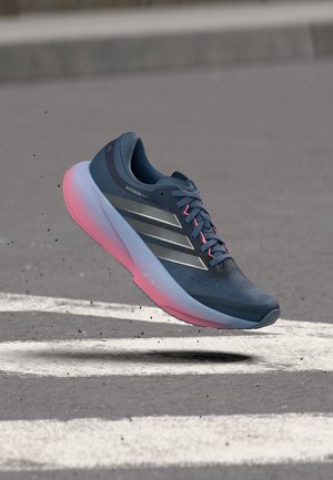 Marineblå Adidas løbesko med lyserøde accenter på sålen og snørebåndene, svævende over asfalt markeret med hvide striber.