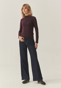 Dlouhý rukáv bordó košile v kombinaci se širokými tmavomodrými džíny. Model má na sobě tmavě hnědé boty a stojí na neutrálním pozadí.