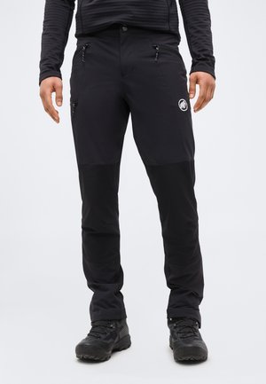 Homme portant un pantalon d'extérieur noir avec poches zippées et des chaussures de randonnée noires, se tenant contre un fond clair uni.