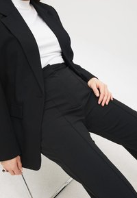 Schwarzes, tailliertes Blazer über einem weißen Rollkragenpullover, kombiniert mit hochgeschnittenen schwarzen Hosen. Glatte Textur und strukturierte Passform. Arm ruht auf dem Oberschenkel.