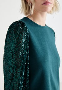 Sudadera azul verdoso con mangas negras de lentejuelas que presentan un patrón texturizado. Tejido suave con un escote redondeado.