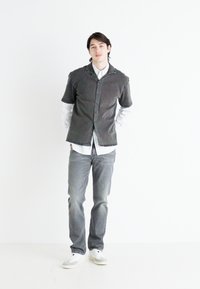 Chemise gris à manches courtes boutonnée par-dessus une chemise blanche à manches longues, associée à un jeans gris et des baskets blanches. Design simple, coupe moderne.
