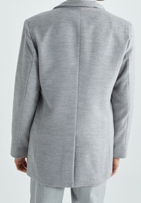 Blazer de lana gris con corte recto y solapas con muescas. Presenta una abertura trasera única y mangas largas. La textura parece lisa y confeccionada a medida.