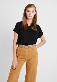 Schwarzes T-Shirt mit kurzen Ärmeln, kombiniert mit hochtaillierten, weiten, senfgelben Hosen und einem hellgrauen Gürtel mit Schnalle und Löchern.