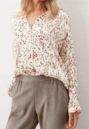 Vrouw draagt een wit bloesje met een bruin gevlekt patroon, ingestopt in lichtbruine hoge taillebroek, staand tegen een effen achtergrond.