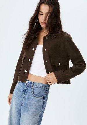 Braune kurze Jacke aus weichem Stoff, mit silbernen Druckknöpfen, einem Kragen und vorderen Taschen. Getragen mit einem weißen Oberteil und blauen Jeans.