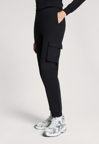Czarne spodnie cargo z miękkiego materiału, z bocznymi kieszeniami, zwężanymi mankietami i gładką fakturą. W połączeniu z srebrnymi butami sportowymi.