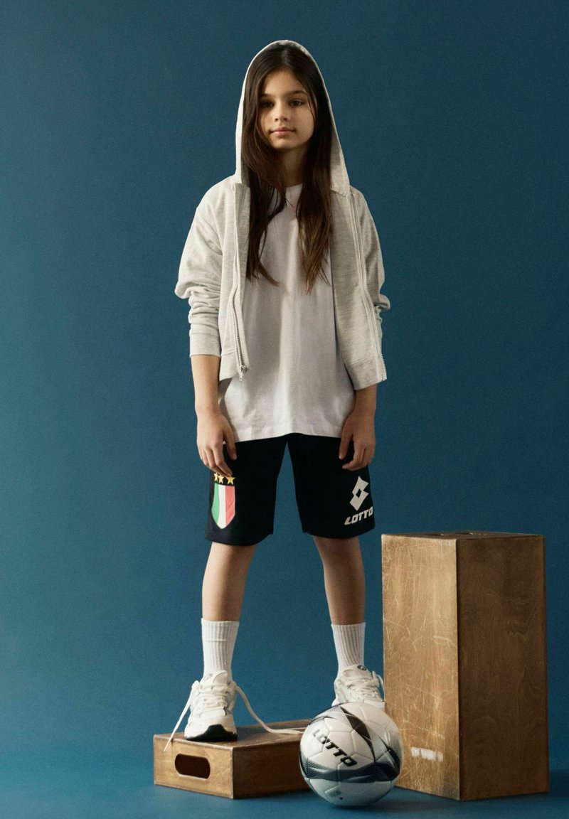 Jeune personne en sweat à capuche gris, short noir et baskets blanches, debout sur des caisses en bois avec un ballon de football sur un fond bleu.