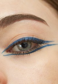 Närbild av ett öga med djärv blå eyeliner och en skarp vinge, långa svarta ögonfransar och en lätt texturerad hudytan.