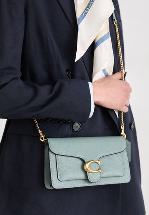 Personne en blazer bleu marine tenant un sac à main en cuir vert clair avec une bandoulière chaîne dorée et une fermeture, portant une écharpe blanche à rayures bleues.