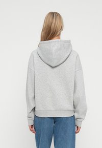 Persona con lunghi capelli biondi che indossa una felpa con cappuccio grigio chiaro e jeans blu, vista di spalle su uno sfondo neutro.