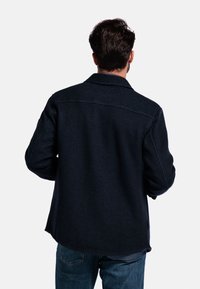 Man in een donkerblauwe jas en blauwe jeans, gezien vanaf de achterkant tegen een effen witte achtergrond.