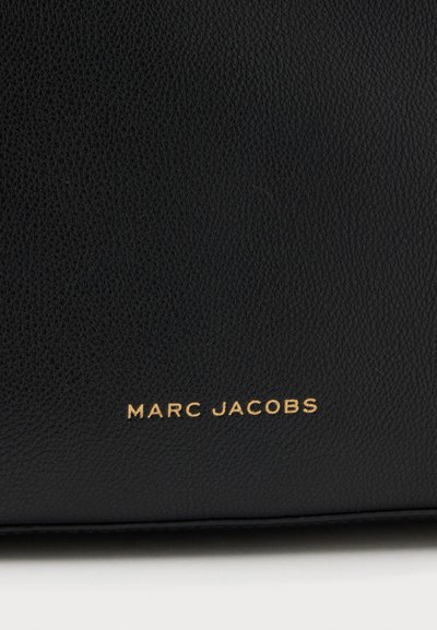 Surface en cuir noir texturé avec texte embossé doré indiquant « "MARC JACOBS" » près du bord inférieur.