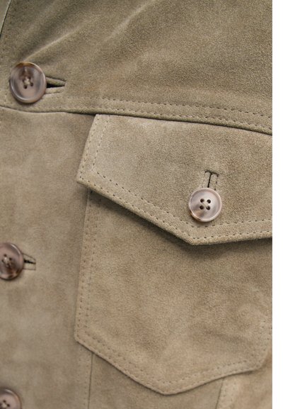 Κοντινό πλάνο από μπεζ σουέτ σακάκι που διαθέτει δύο κουμπιά, μια τσέπη στο στήθος με καπάκι και λεία υφή με ορατές ραφές.