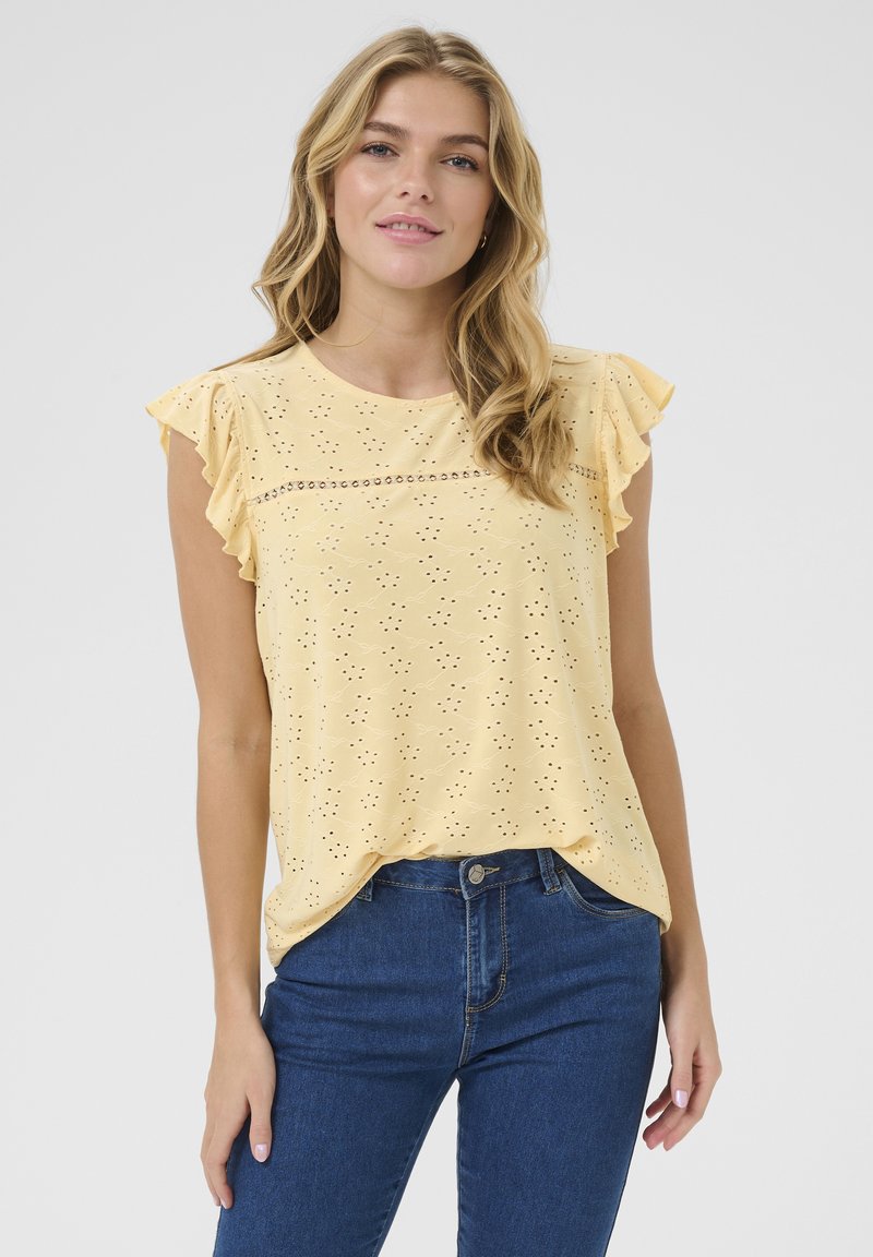 Haut jaune clair avec des manches froncées, motif en oeillets et une couture décorative horizontale. Associe à un jean bleu foncé. Texture lisse.