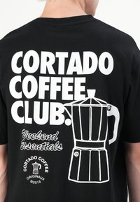 Schwarzes Baumwoll-T-Shirt mit auffälligem weißen Text: "CORTADO COFFEE, CLUB." Enthält eine Grafik einer Kaffeemaschine und die Schrift "Weekend Essentials".