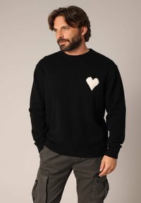 Zwarte trui van zacht materiaal, met een geborduurd wit hartmotief op de linkerborst. Ribbelkraag en -manchetten.